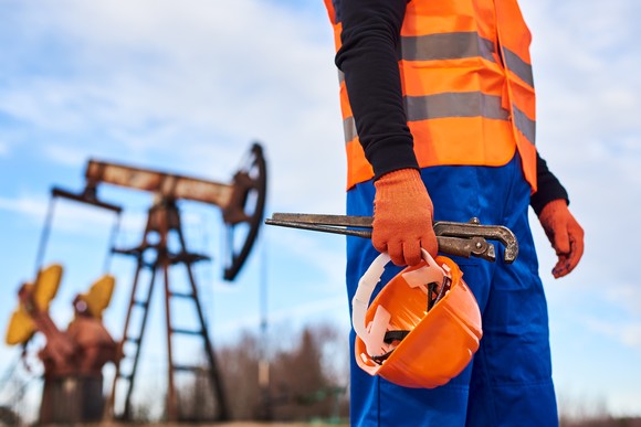 Oil field services worker holds helmet in front of a rig. 