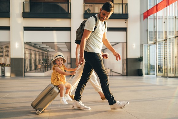 Two adults walk through an airport. A child rides on the handle of a wheeled suitcase.