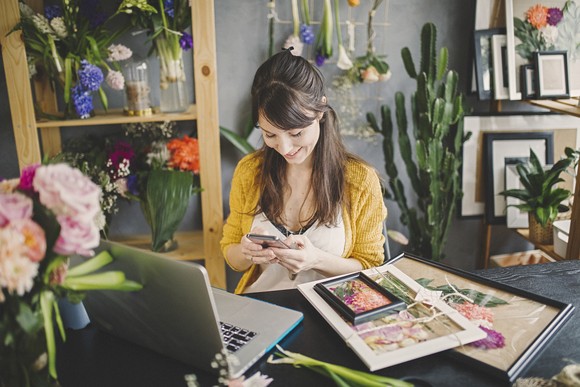 A person sitting in front of a computer with flowers and other products for sale surrounding them.
