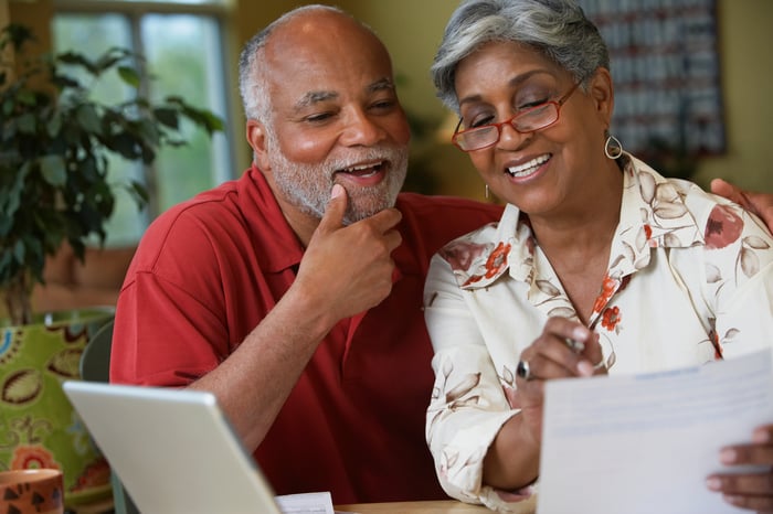 A smiling couple reviews paperwork in front of a laptop at home.