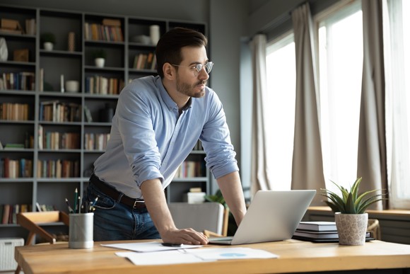 An investor stands over a desk with a laptop on it and gazes out a window.