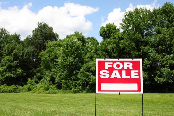 For sale sign in front of vacant land with grass and trees.
