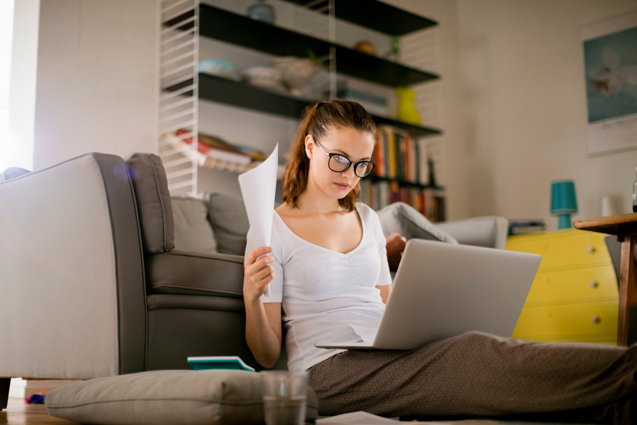 Person wearing glasses sits on floor holding paperwork and looking at a laptop screen. 