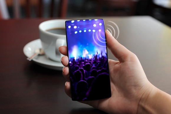 A hand holds a smartphone with the thumb pressing a haptic-enabled button on the side. In the background is a wooden table with a coffee cup on it