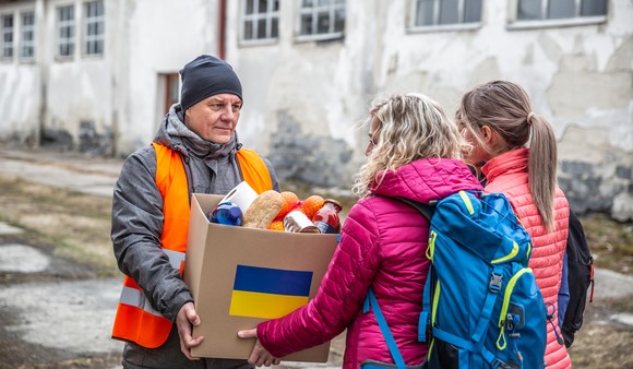 A person wearing an orange vest gives a cardboard box with a Ukraine flag on it to two people.