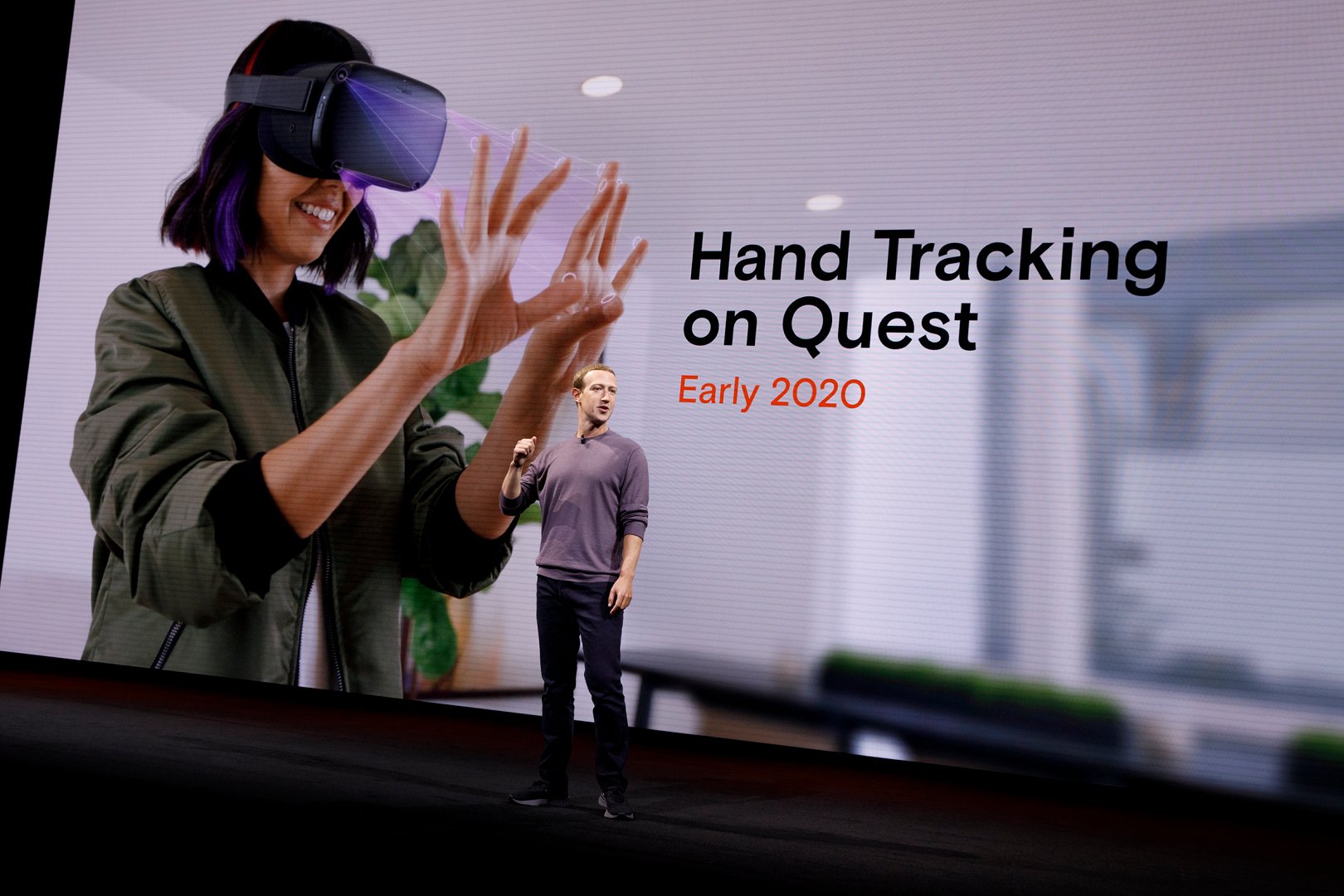 Meta Platforms CEO Mark Zuckerberg stands in front of a screen showing a person wearing an Occulus Quest device and looking at the person's hands