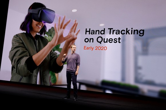 Meta Platforms CEO Mark Zuckerberg stands in front of a screen showing a person wearing an Occulus Quest device and looking at the person's hands