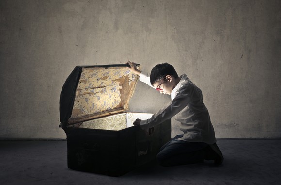 A young person opens a chest and looks down at something gleaming inside.