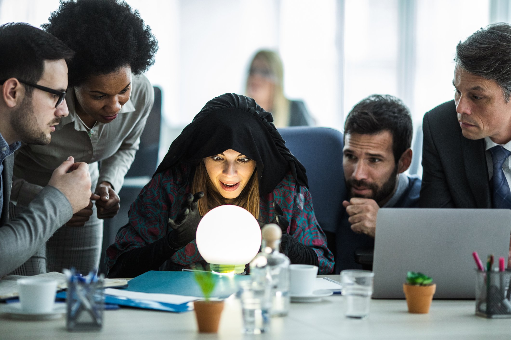 Group of professionals in an office gathered around a cloaked person looking into a crystal ball.