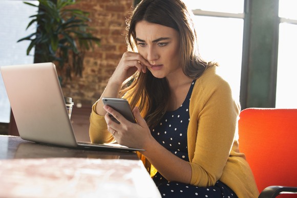 Person with a worried expression sitting at a desk and staring at a smartphone.