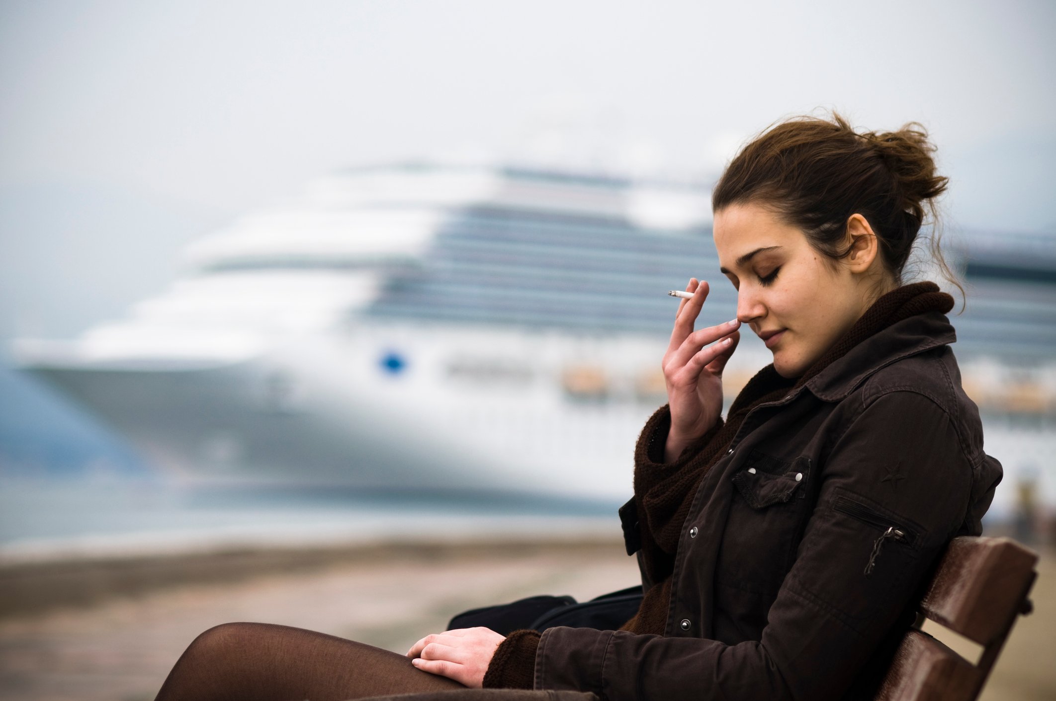 Displeased person smoking a cigarette while seated in a chair, with a cruise ship in the background.