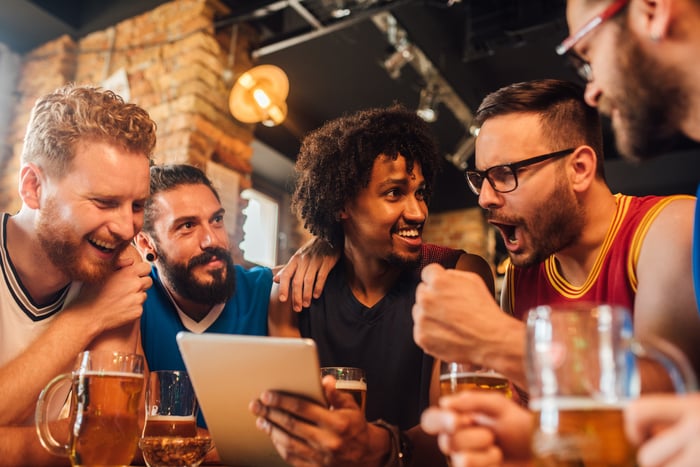 A group of young people gathered at a table, reacting excitedly to something on a tablet computer.