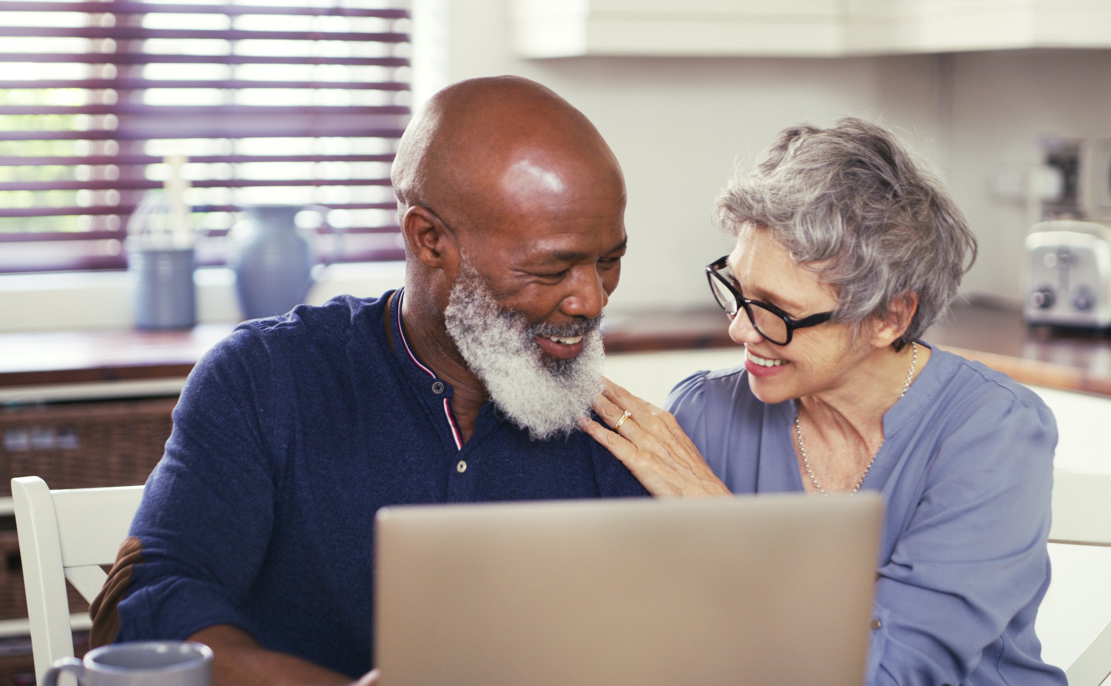 Smiling senior couple sitting in kitchen using laptop.
