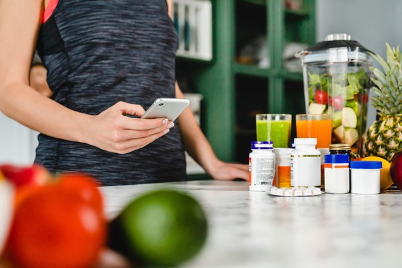 Person with phone standing near a table with nutritional supplements and fruit.