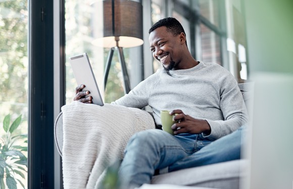 A person smiles while looking at something on a tablet while sitting on a couch.