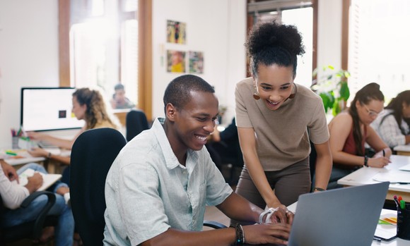 Two workers smile while looking at something on a laptop at work.