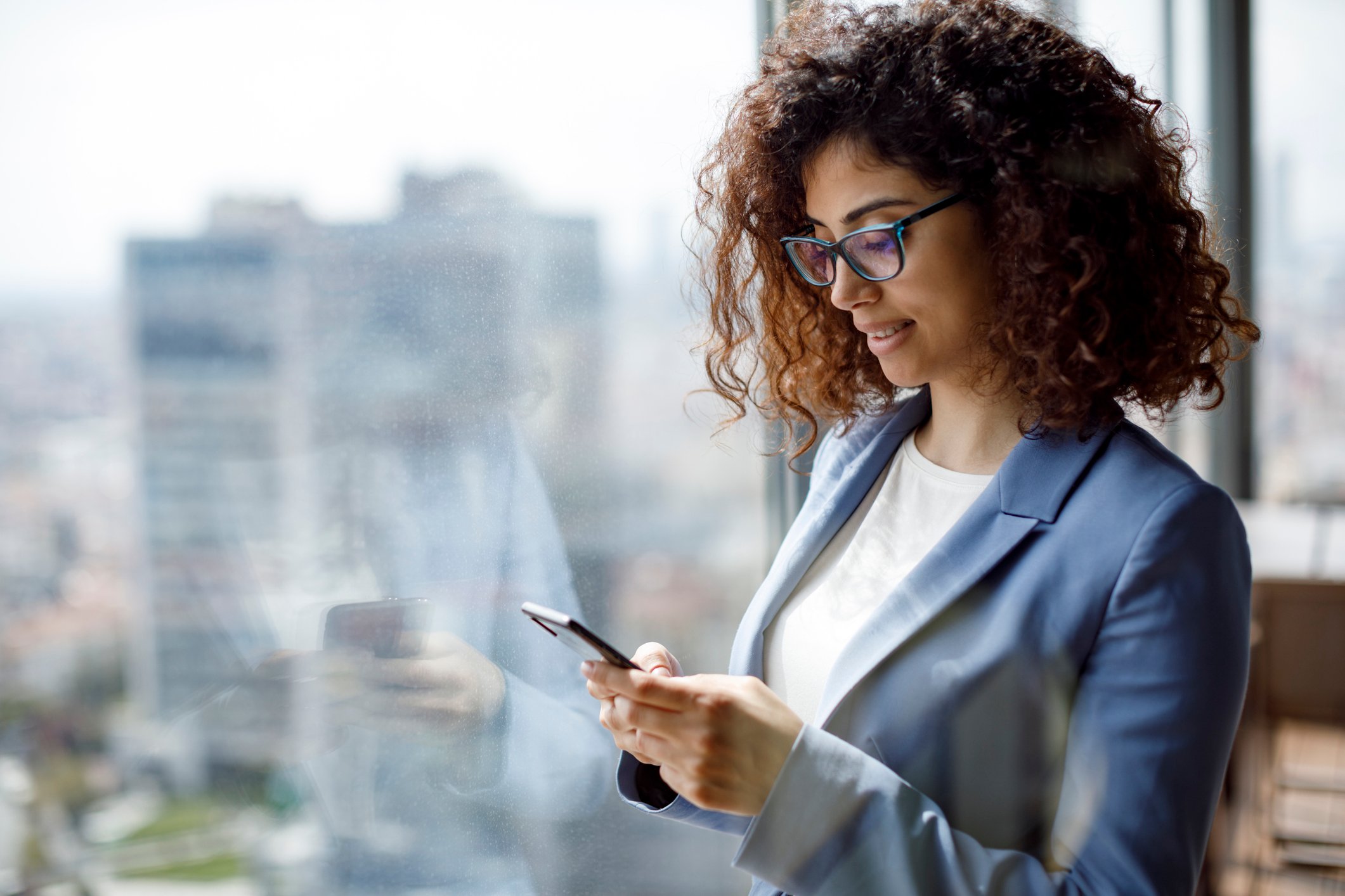 Office worker checks smartphone while standing by a window in a high-rise office complex.