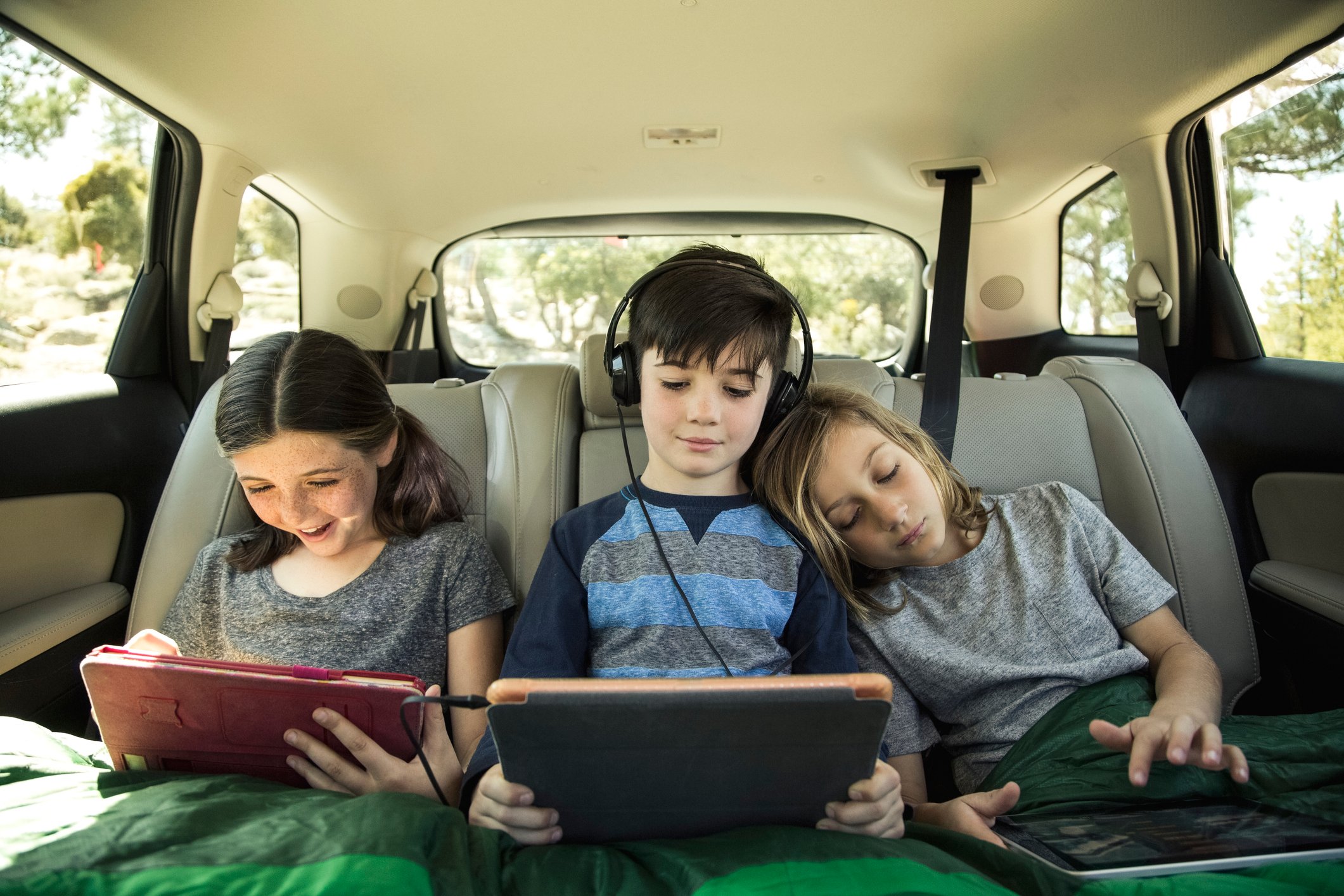 Three children using tablets in the backseat of a car.