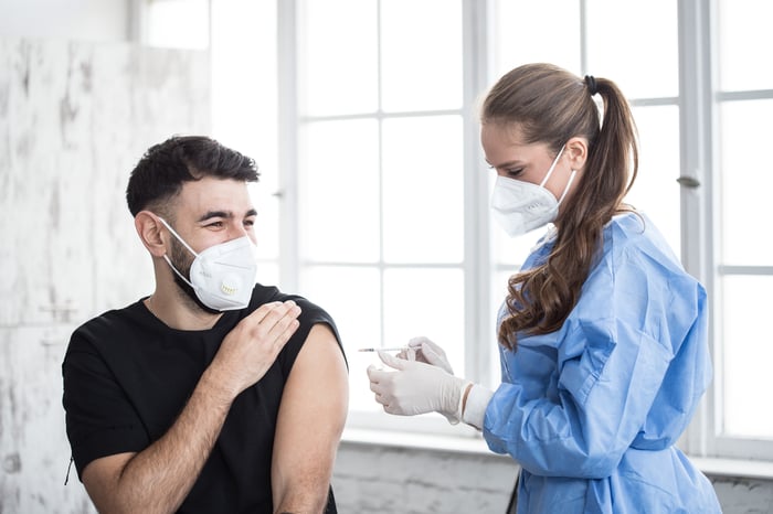 A nurse vaccinates a person in a clinic while wearing protective equipment.