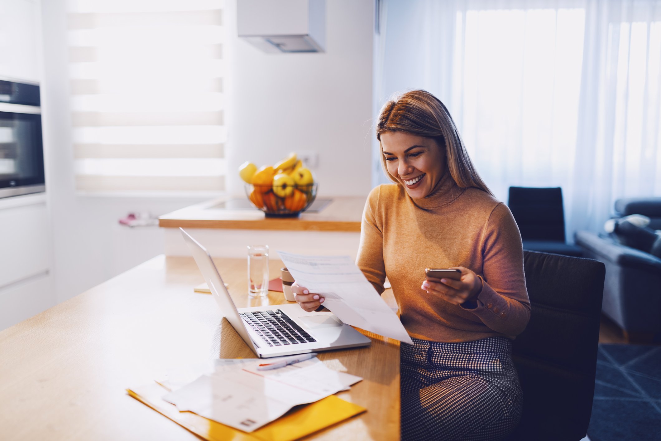 Person looking at paper smiling while holding phone in front of laptop.