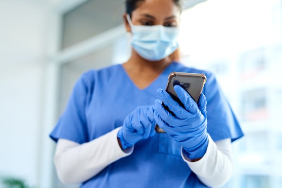 A person in medical scrubs, mask, and gloves holding a smartphone.