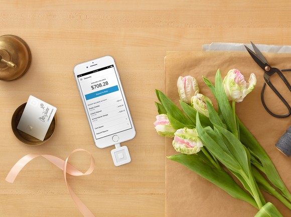 A Square card reader plugged into a smart phone on a counter with flowers.