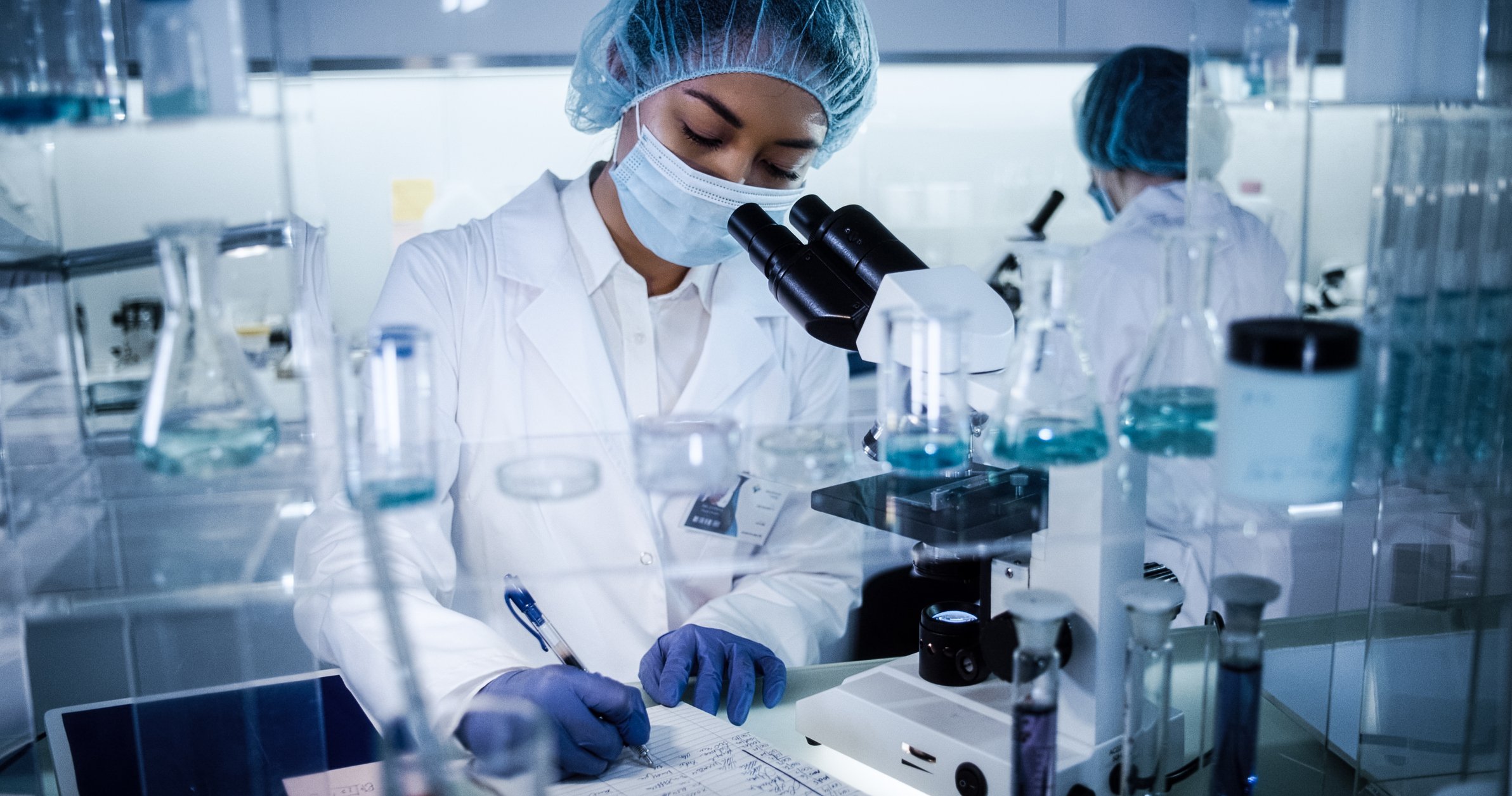 A scientist writing on a document while standing in front of a microscope in a lab.