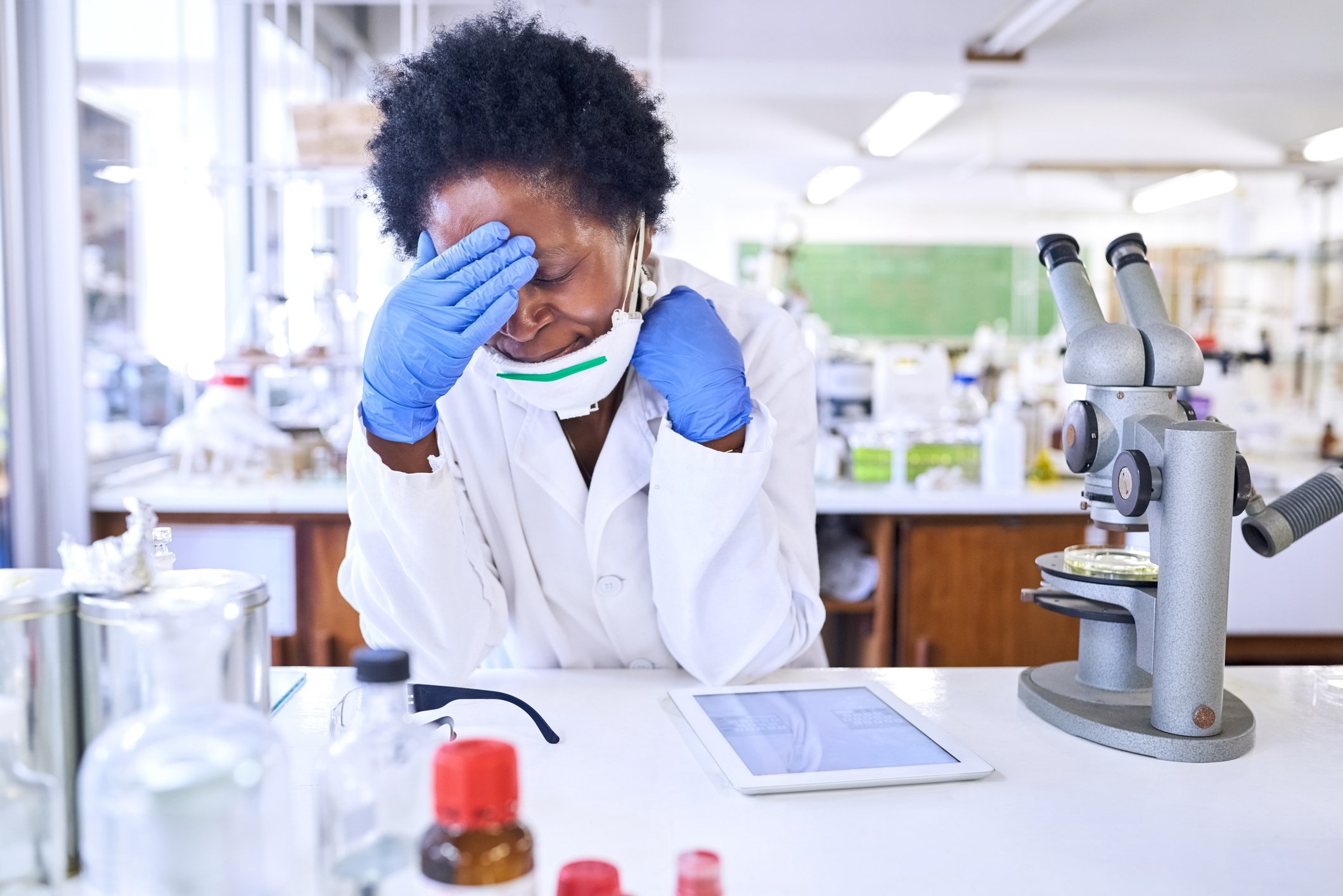 A scientist touches head in exasperation while sitting at a laboratory bench with a tablet and a microscope.