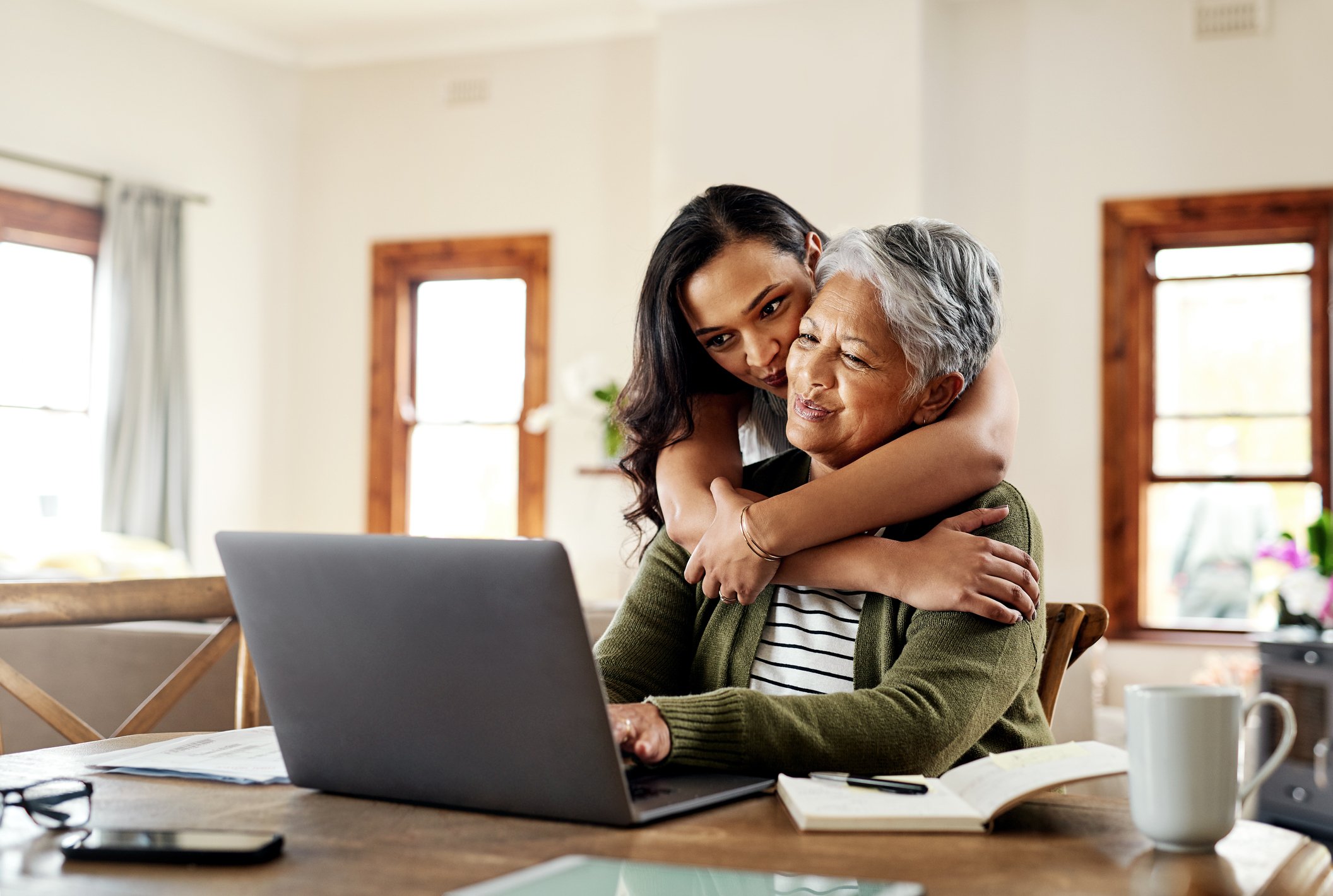 A lady hugs an older lady who is smiling while working on a computer.