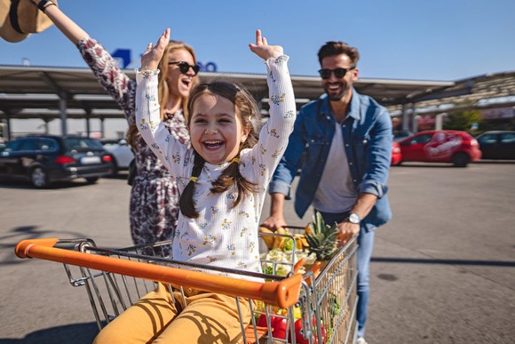 A child throws their hands up in glee as two adults push them in a shopping cart through a parking lot. 