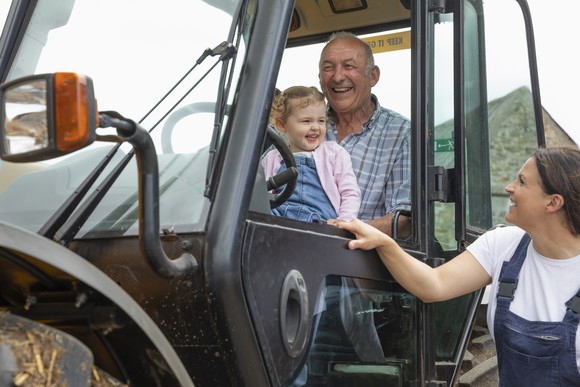 An older person sits in the cab of a tractor with a child. Both smile as a third person outside the tractor also smiles at the child.