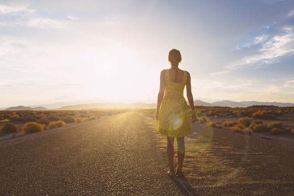 A person stands on an open remote road facing the rising sun.