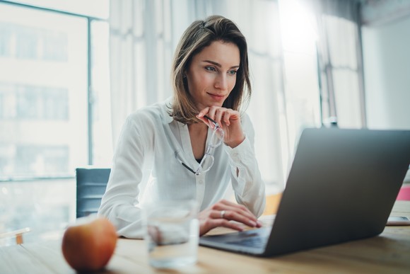 A professionally dressed individual looks thoughtfully at a laptop screen, with a glass of water and an apple nearby.