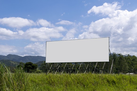 Blank billboard in field with blue sky and mountains in background.
