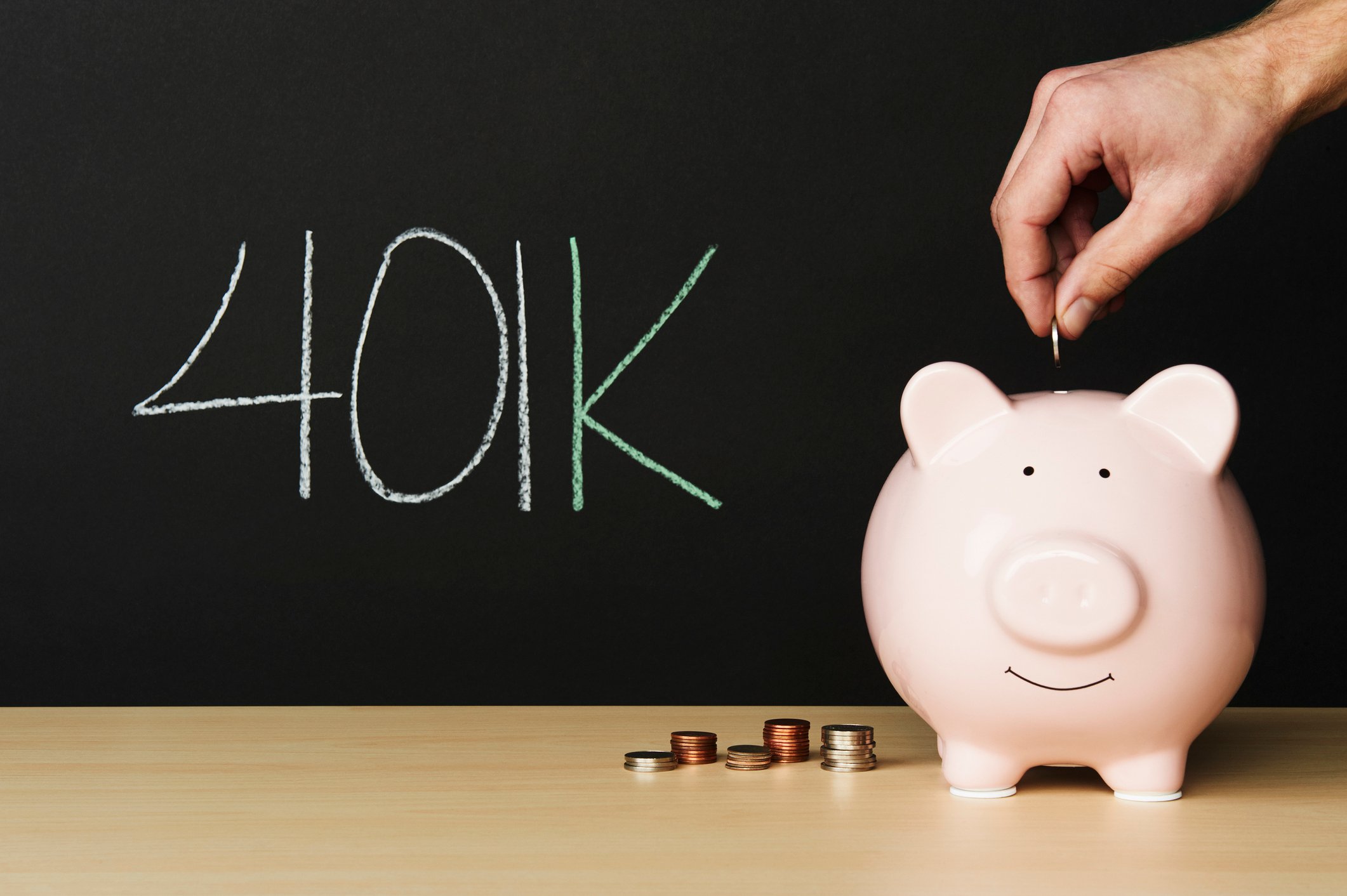 Piggy bank with a smiling face on a desk with coins and 401(k) written on a chalk board.