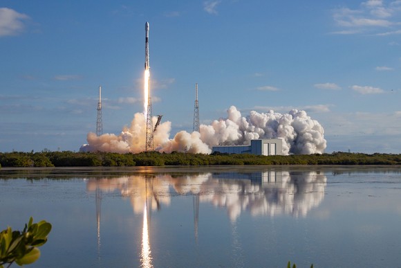 A SpaceX Starlink 2 rocket takes off with water in the foreground and blue skies in the background