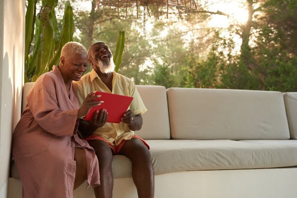 Two people sit on couch in front of window, smiling and holding a laptop. 