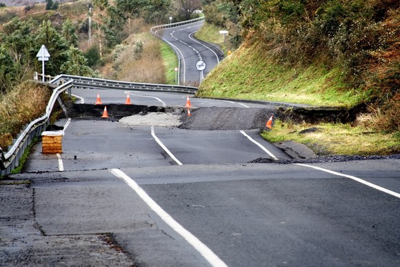 An uneven paved road with potholes and broken concrete in a rural setting. 