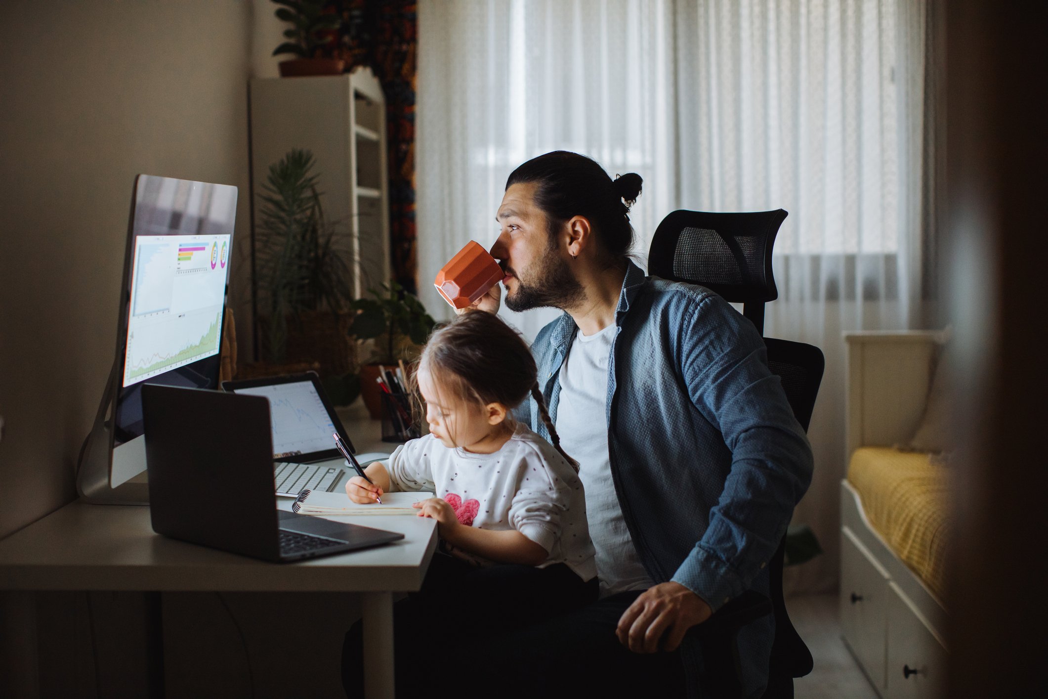 A parent drinks coffee and researches stocks on a computer while holding a child.