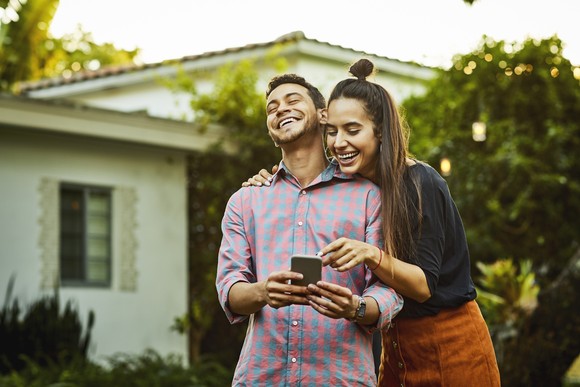 Two people laugh and smile while looking at smartphone and standing in front yard of home.