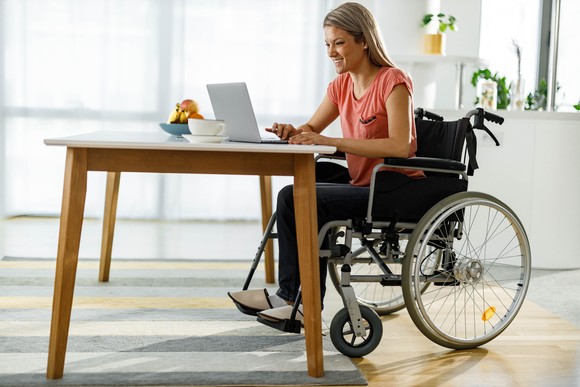 Person smiles while sitting in wheelchair at table with laptop.
