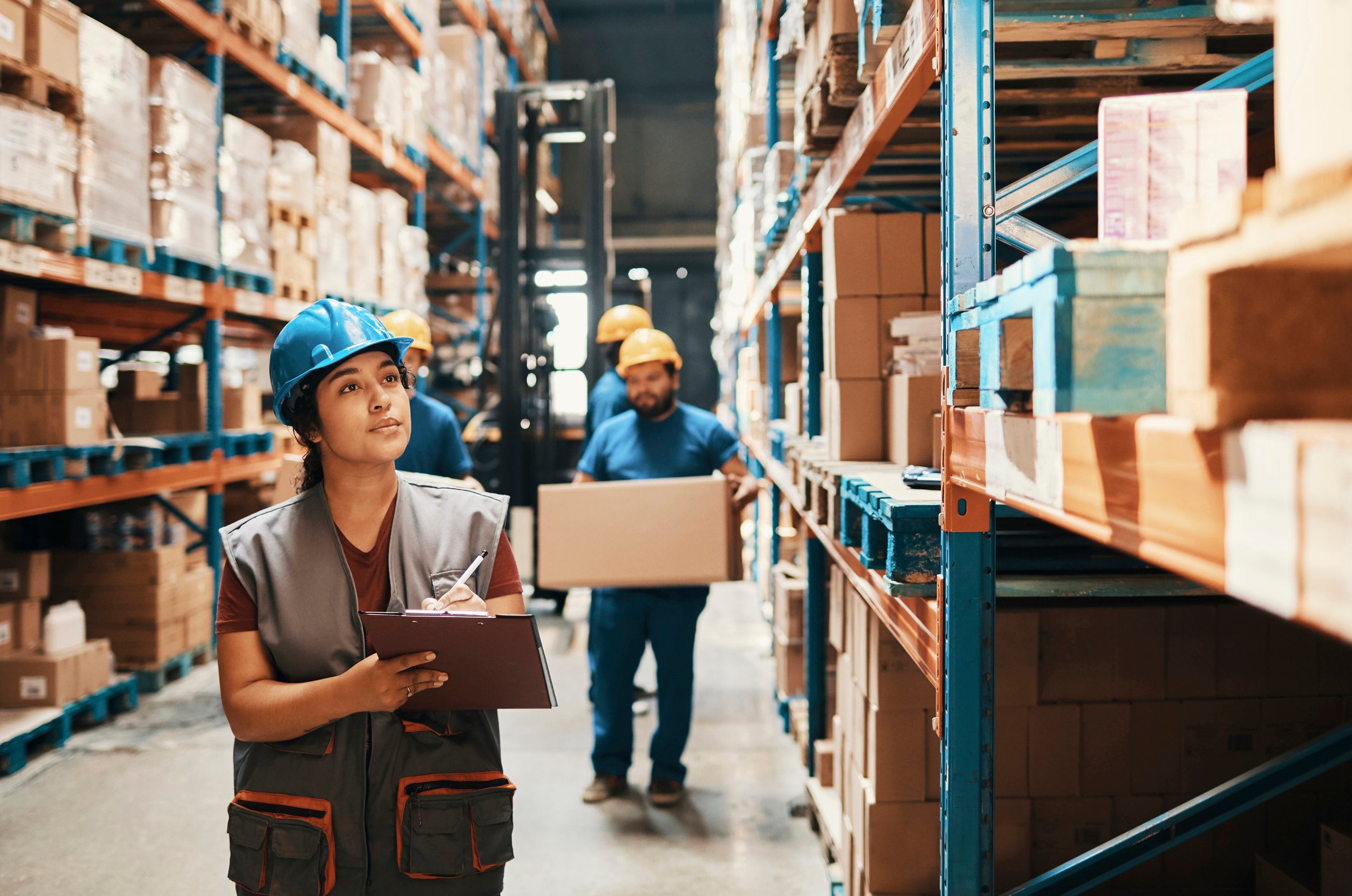 Workers carry packages in a warehouse behind another worker with a clipboard.
