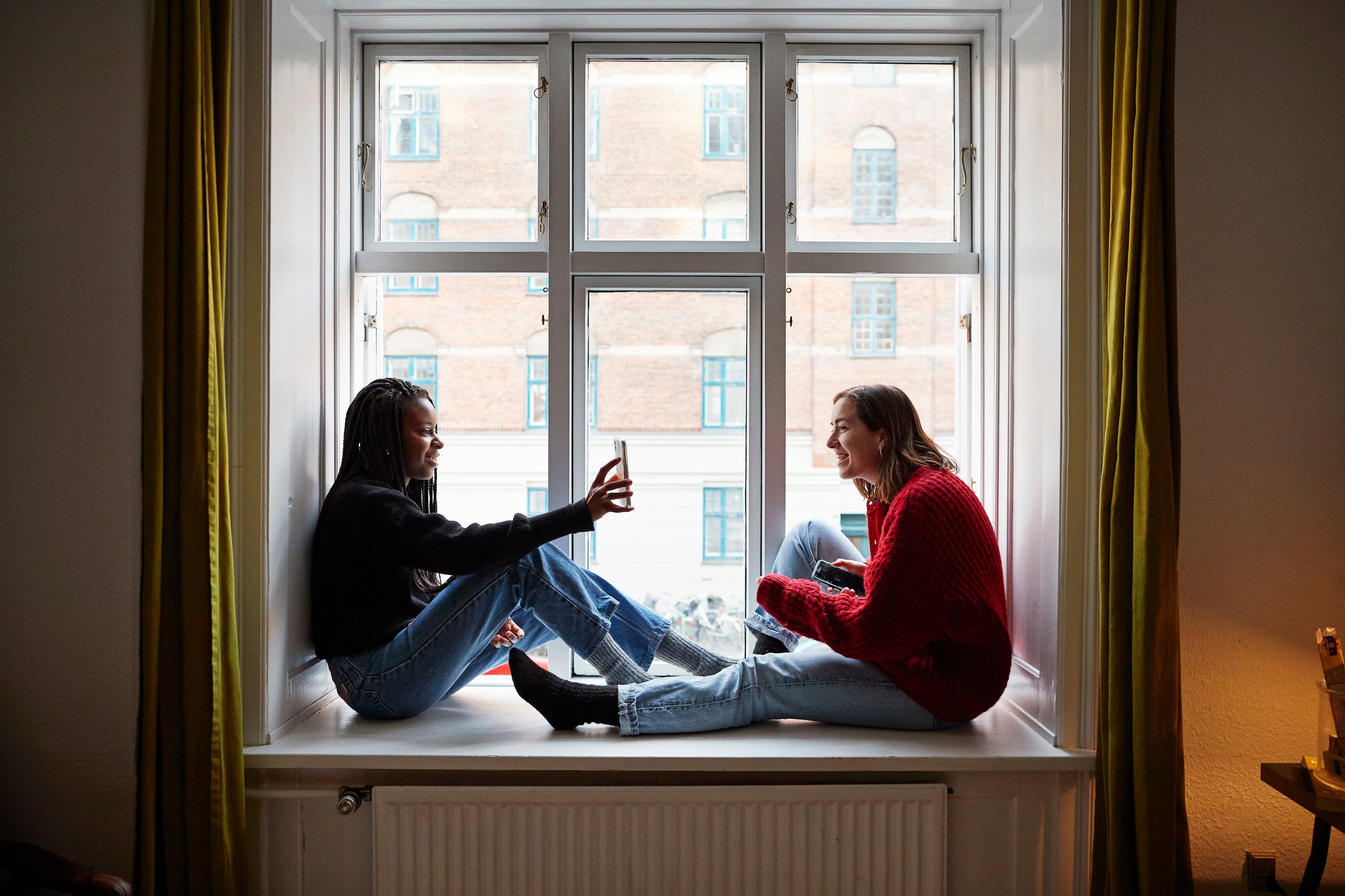 Two people sittng near an apartment window while sharing phone screens by the window.