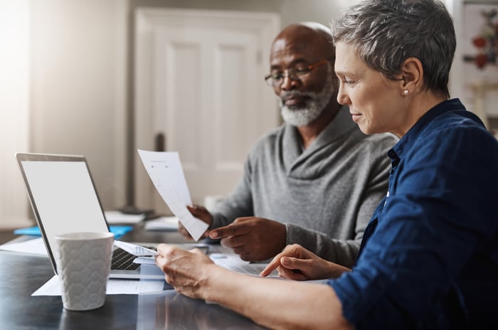 Two people study paperwork and laptop screen while sitting at table together.