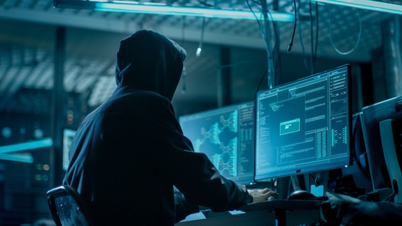A hooded hacker sits at a computer in a dark room.