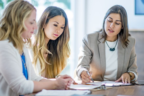 Three investors sit at a table while one gestures to a binder with papers as the others look on.