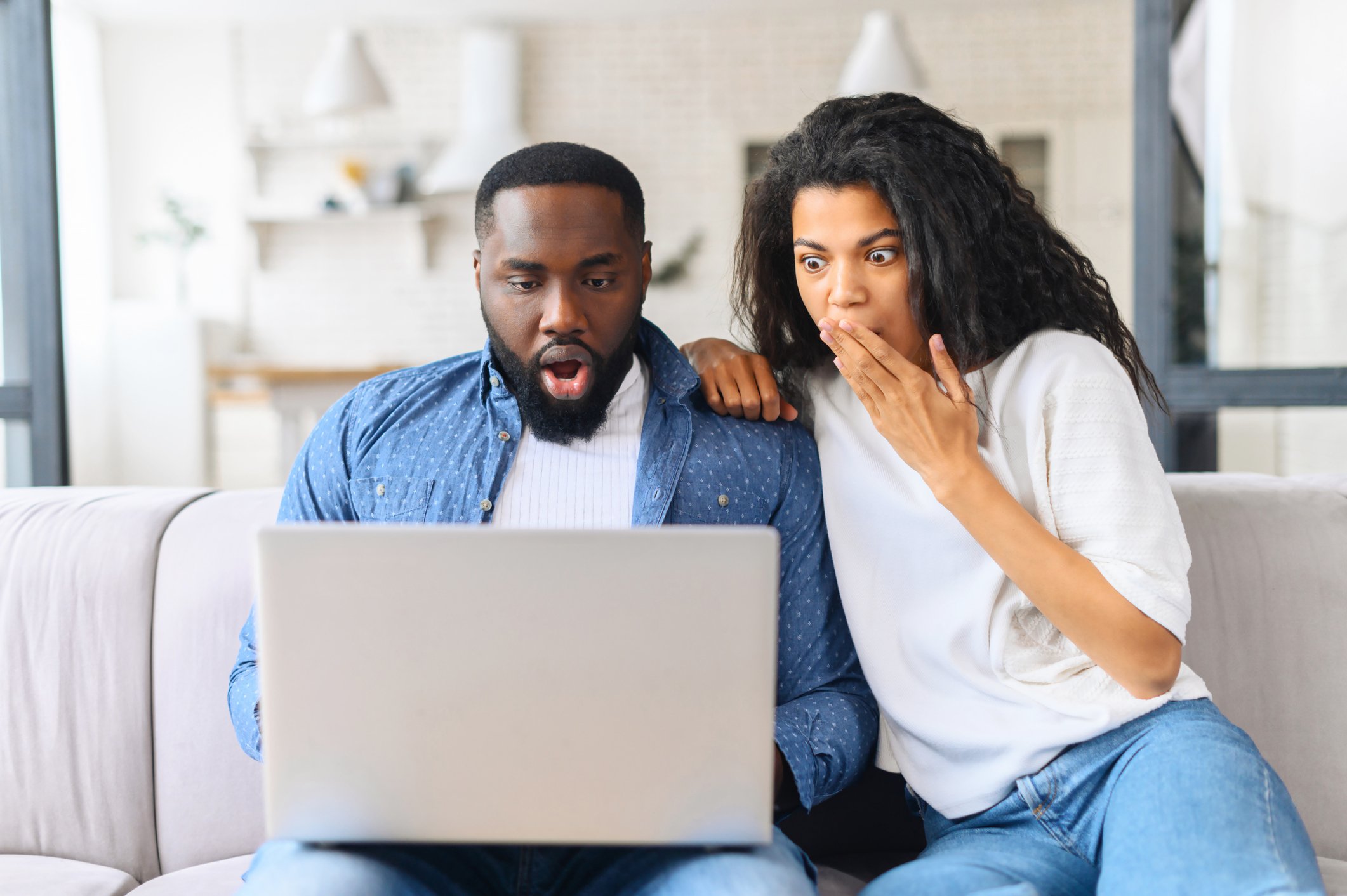 Two people sitting on sofa are looking shocked at laptop screen.