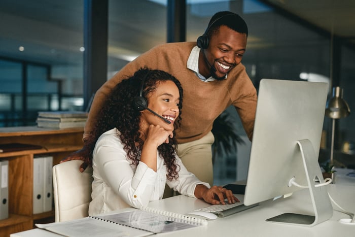 Two workers with headsets interact with others via a desktop.