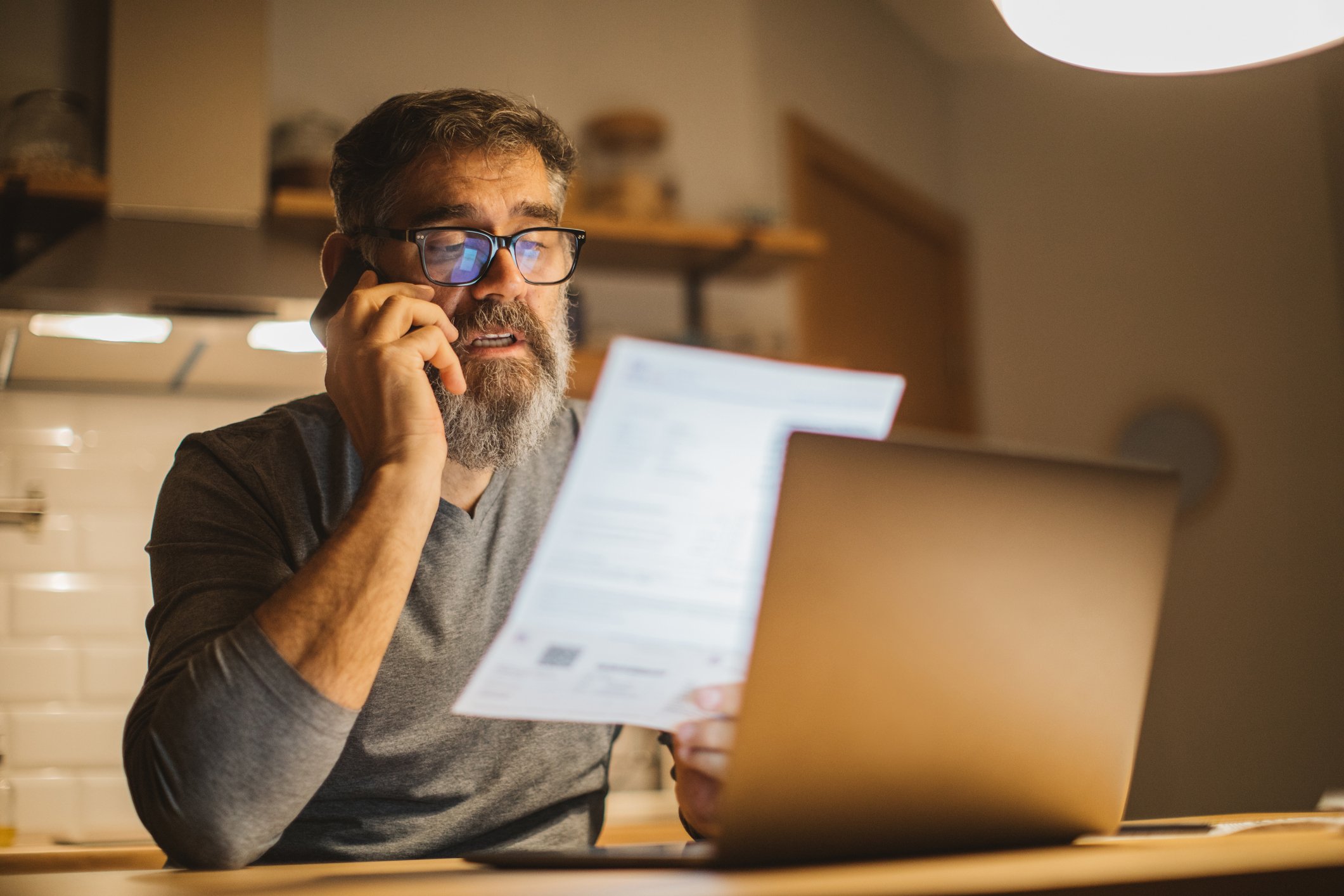 Person wearing glasses talks on phone while holding paperwork and looking at laptop.