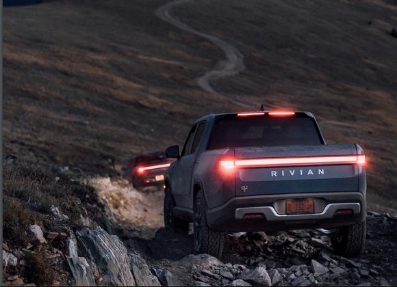 Two Rivian trucks heading up mountain trail at dusk.